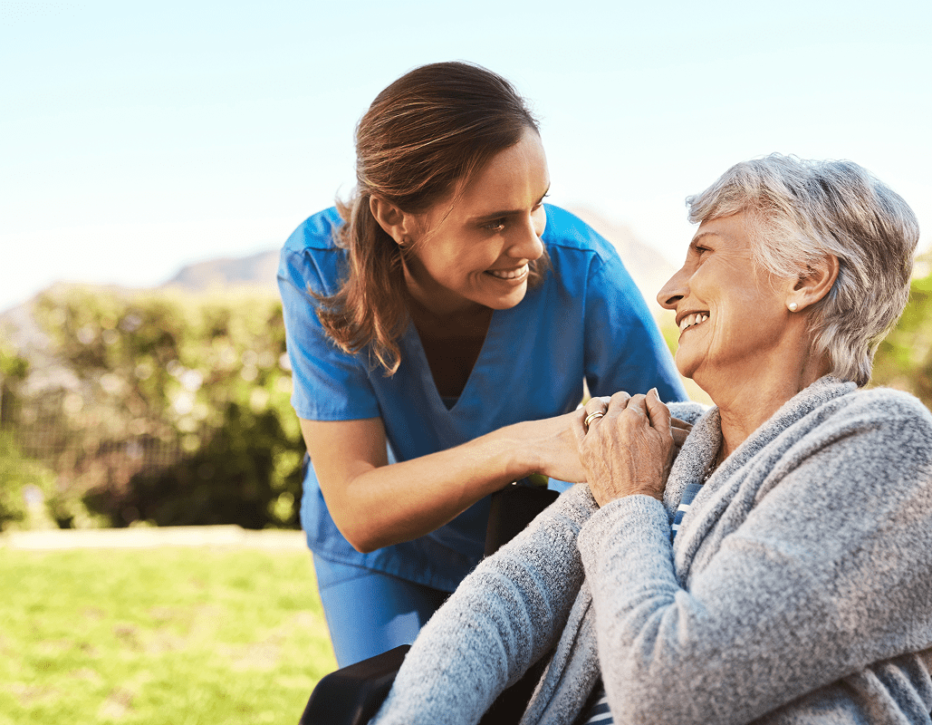 A healthcare worker in blue scrubs smiling and holding hands with an elderly woman sitting outside on a sunny day, both appearing happy and engaged.