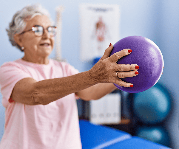 An elderly woman with glasses and gray hair, wearing a pink shirt, holding a purple exercise ball with both hands during a therapy session in a rehabilitation center.