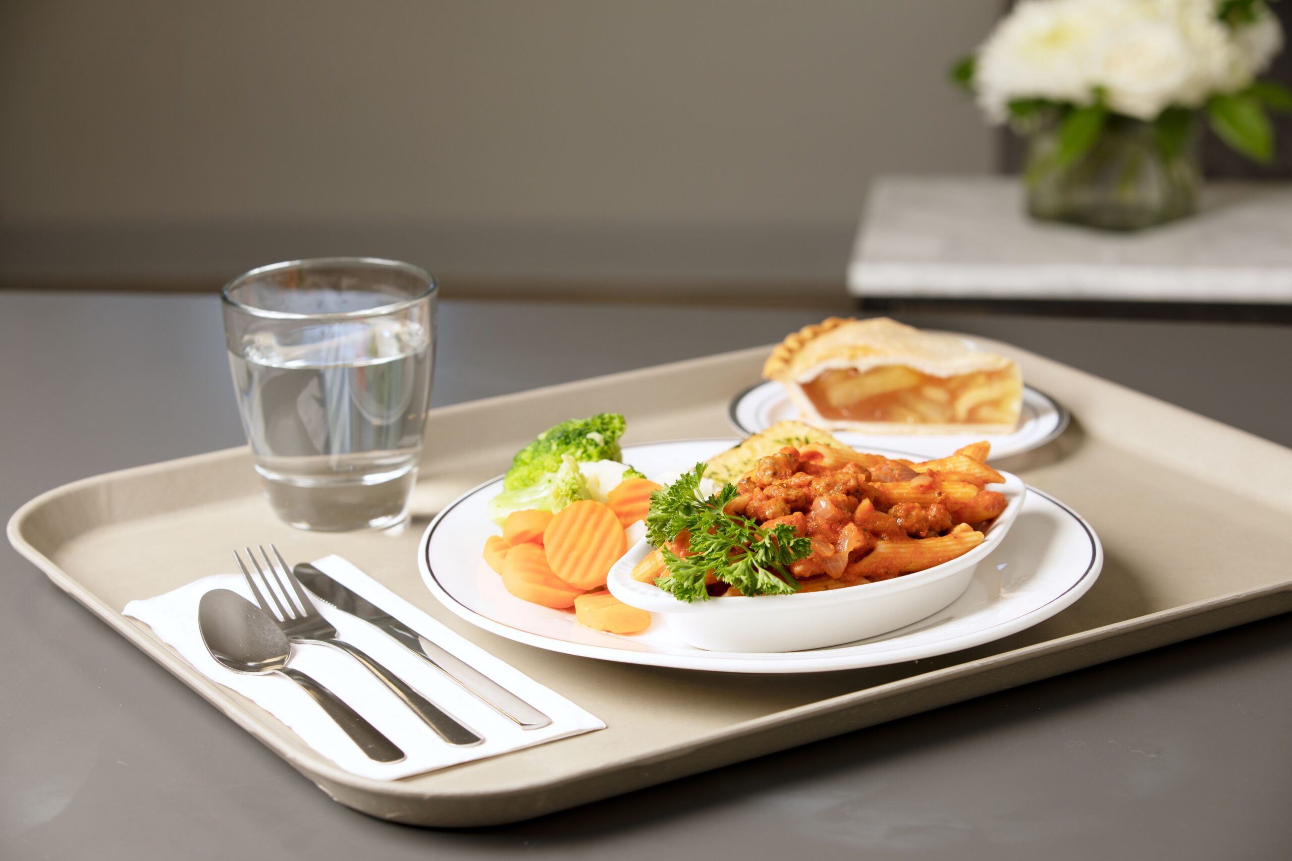 A tray with a glass of water, a plate of mixed vegetables, a bowl of pasta with sauce and garnished with parsley, and a slice of apple pie.